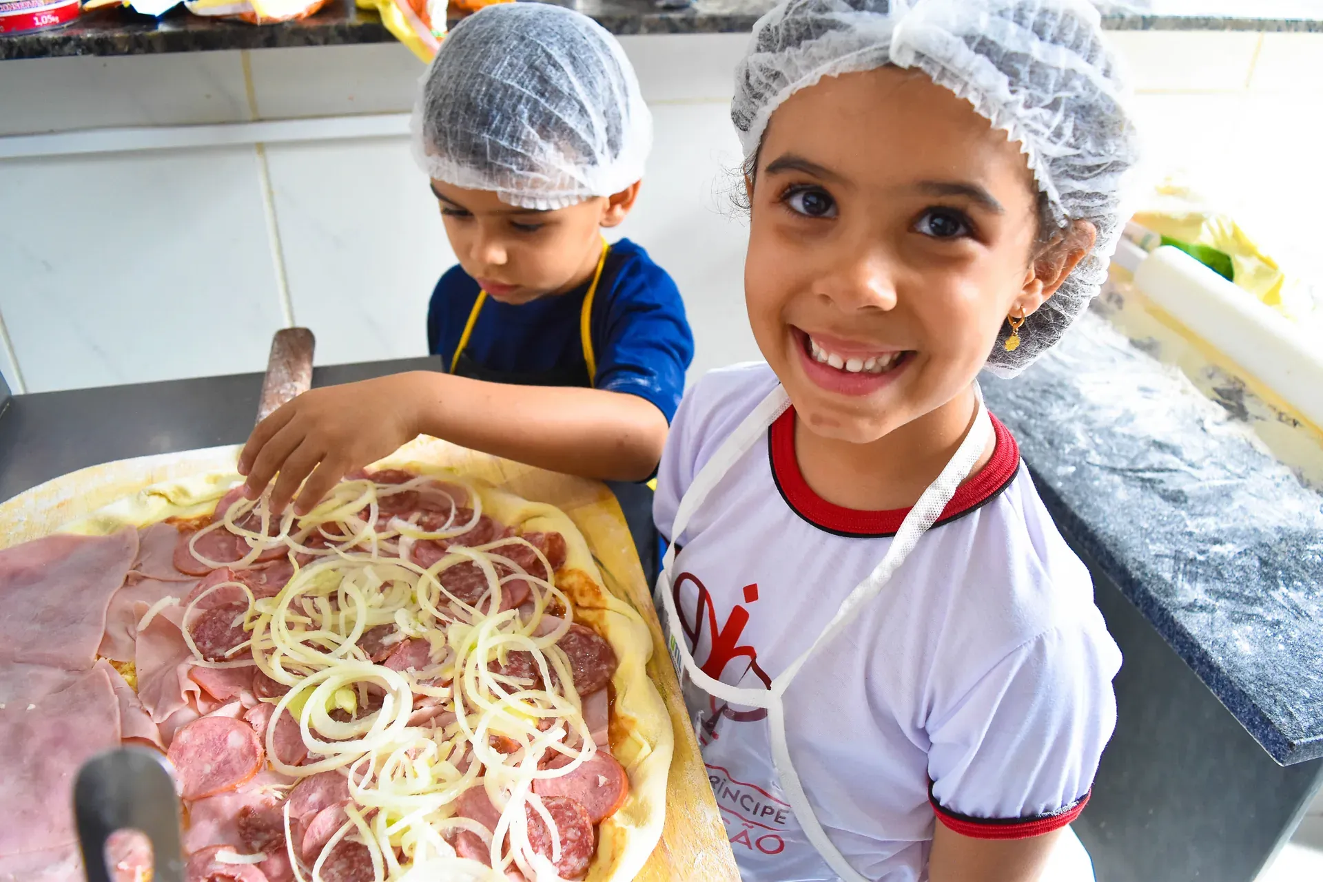 Dupla de alunos de touca e avental, orgulhosos da pizza que acabaram de montar