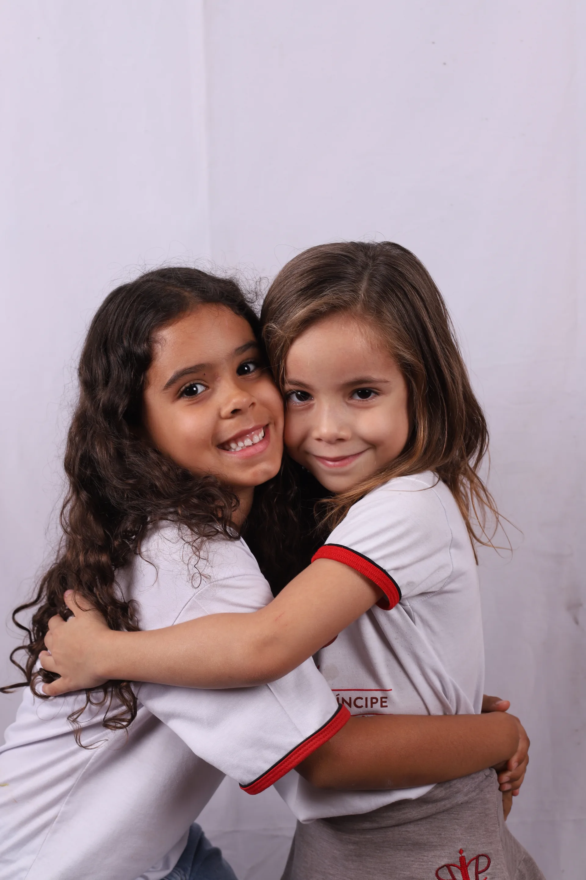 Duas alunas pequenas se abraçam no ensaio em fundo branco da campanha